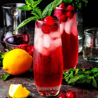 shot of cranberry gin cocktails sitting on a dark surface with a lemon and fresh mint next to the glasses.