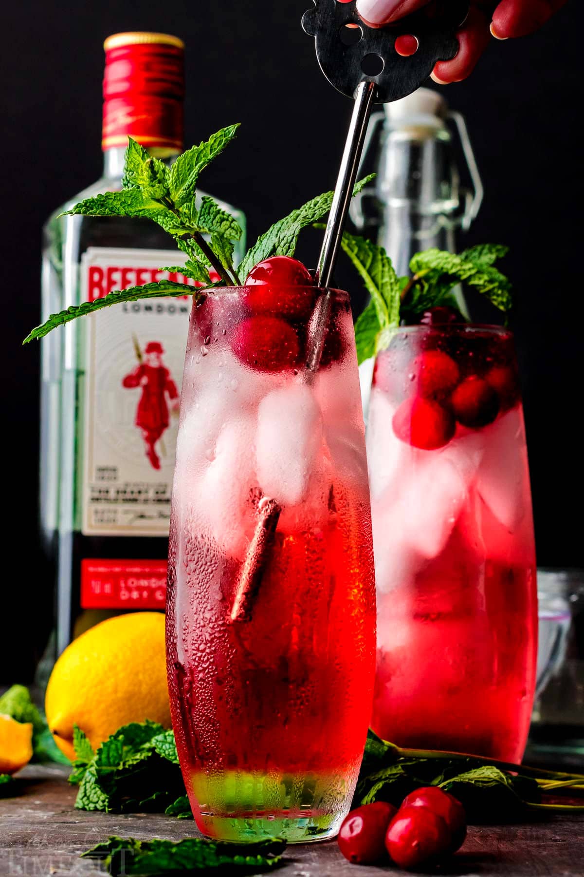 un agitatore di metallo che mescola il cocktail di gin al mirtillo rosso e un altro cocktail è sullo sfondo con una bottiglia di gin.