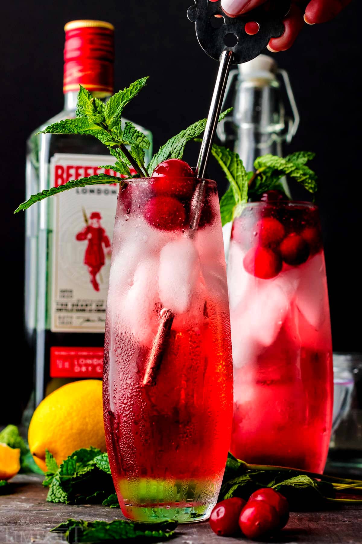 a metal stirrer stirring the cranberry gin cocktail and another cocktail is in the background with a bottle of gin.