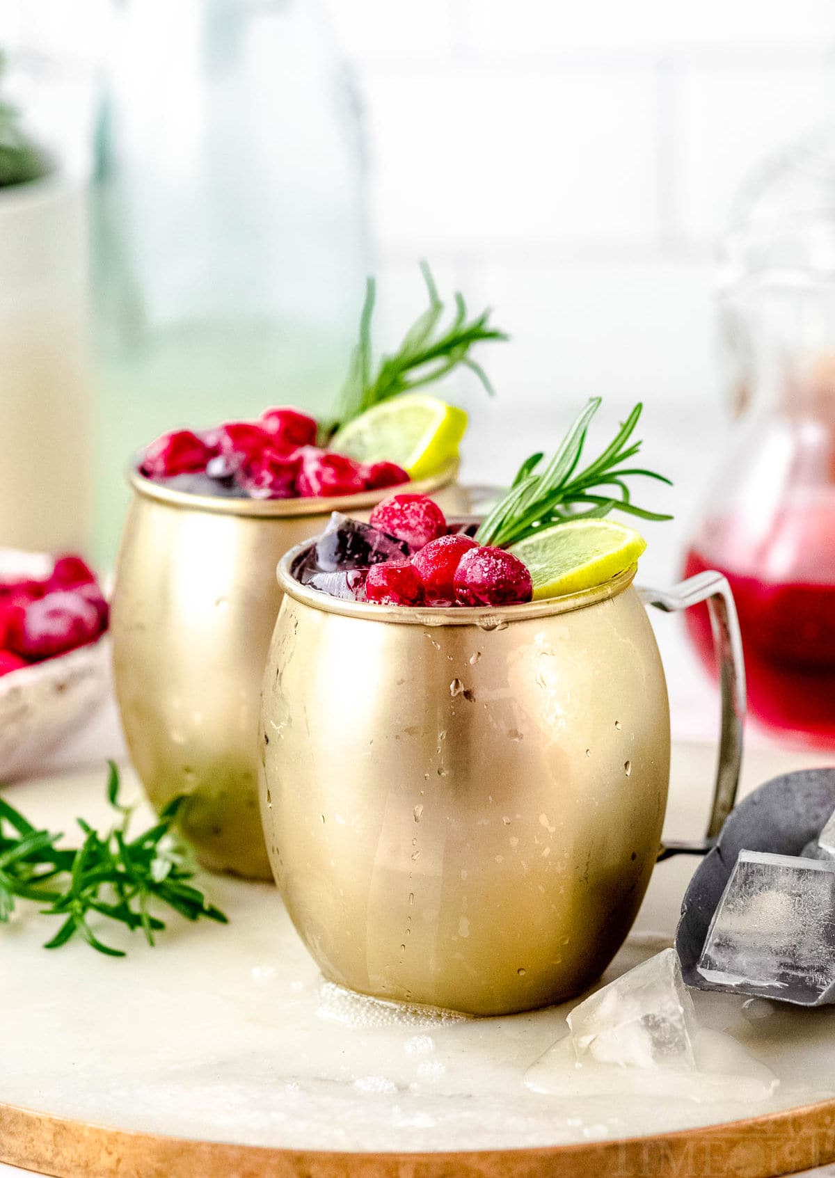 two cranberry moscow mule cocktails on a white board garnished with cranberries, lime slices and a sprig of rosemary.