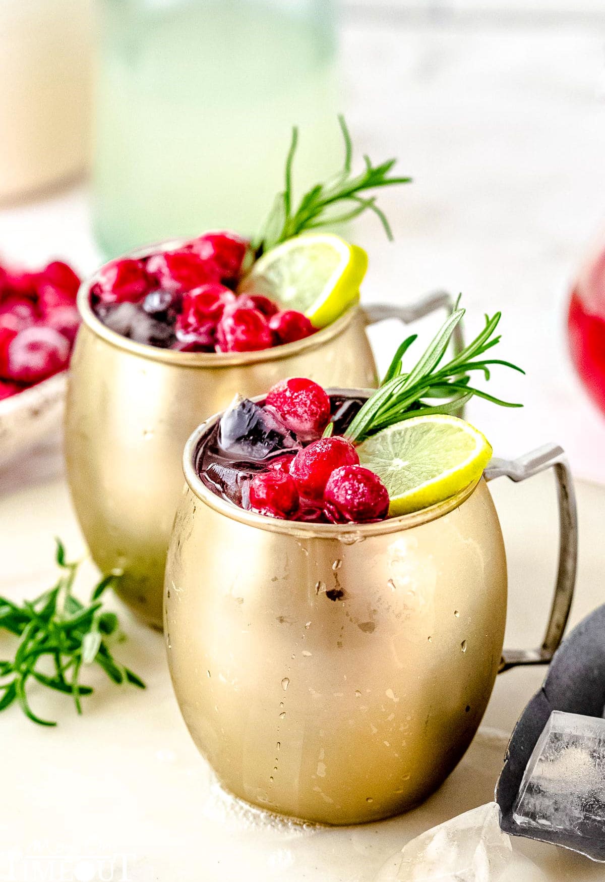 two cranberry moscow mule cocktails on a white board garnished with cranberries, lime slices and a sprig of rosemary.