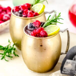 two cranberry moscow mule cocktails on a white board garnished with cranberries, lime slices and a sprig of rosemary.