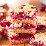 three cranberry crumb bars stacked on each other on a dark wood board with a glass of milk in the background.