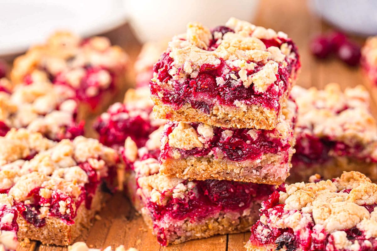 three cranberry crumb bars stacked on each other on a dark wood board with a glass of milk in the background.