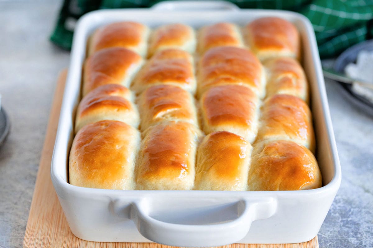 white baking dish with golden brown parker house rolls ready to be enjoyed. baking dish is sitting on cutting board.