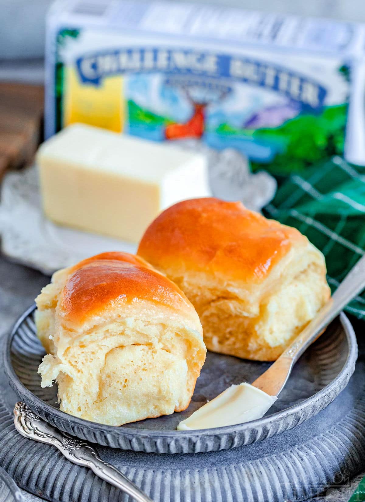 two parker house dinner rolls on a metal plate with a stick of butter behind them. a box of butter can be seen in the background.