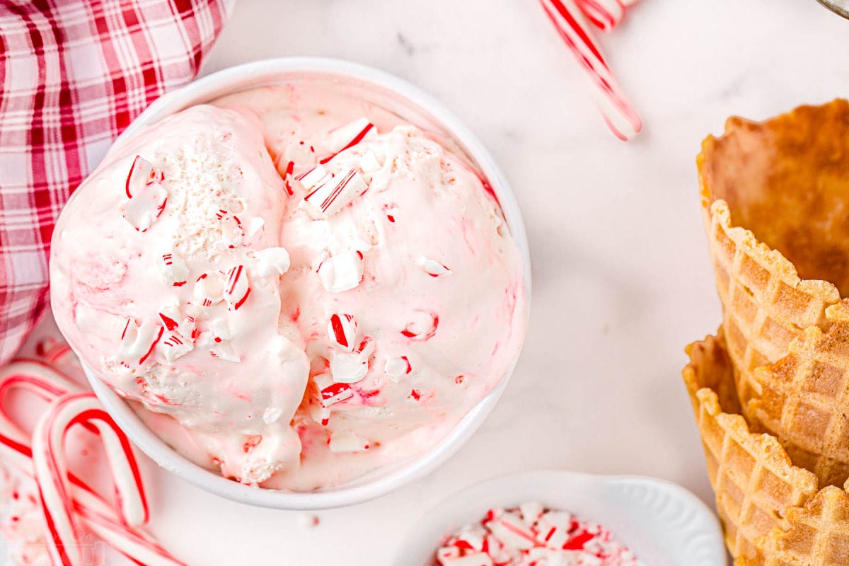 top down view of white bowl filled with peppermint ice cream sitting next to a red checked christmas linen. two waffle cones are sitting near the bowl.