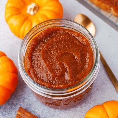 top down look at glass jar filled with pumpkin butter. gold spoon in the pumpkin butter and three small pumpkins surround the jar.