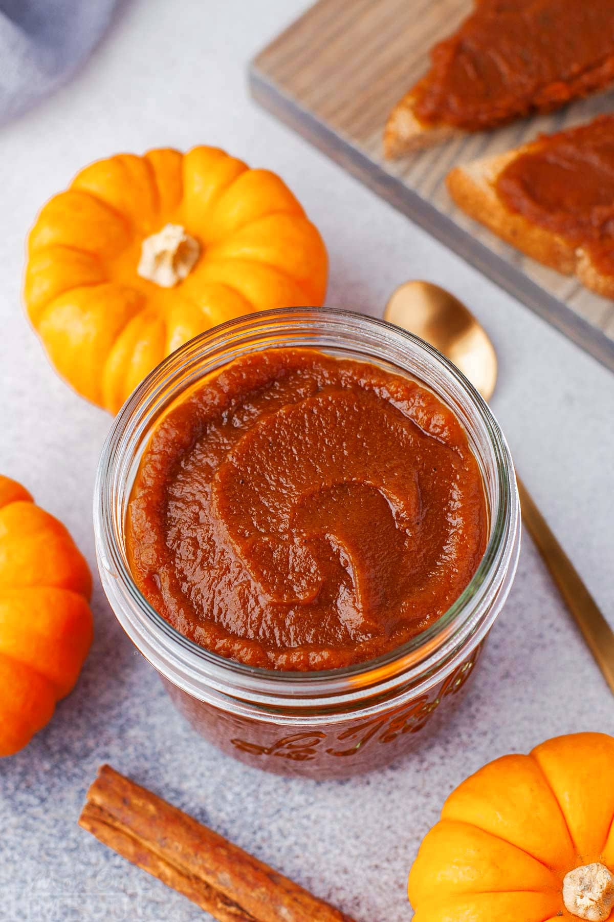 top down look at glass jar filled with pumpkin butter. gold spoon in the pumpkin butter and three small pumpkins surround the jar.