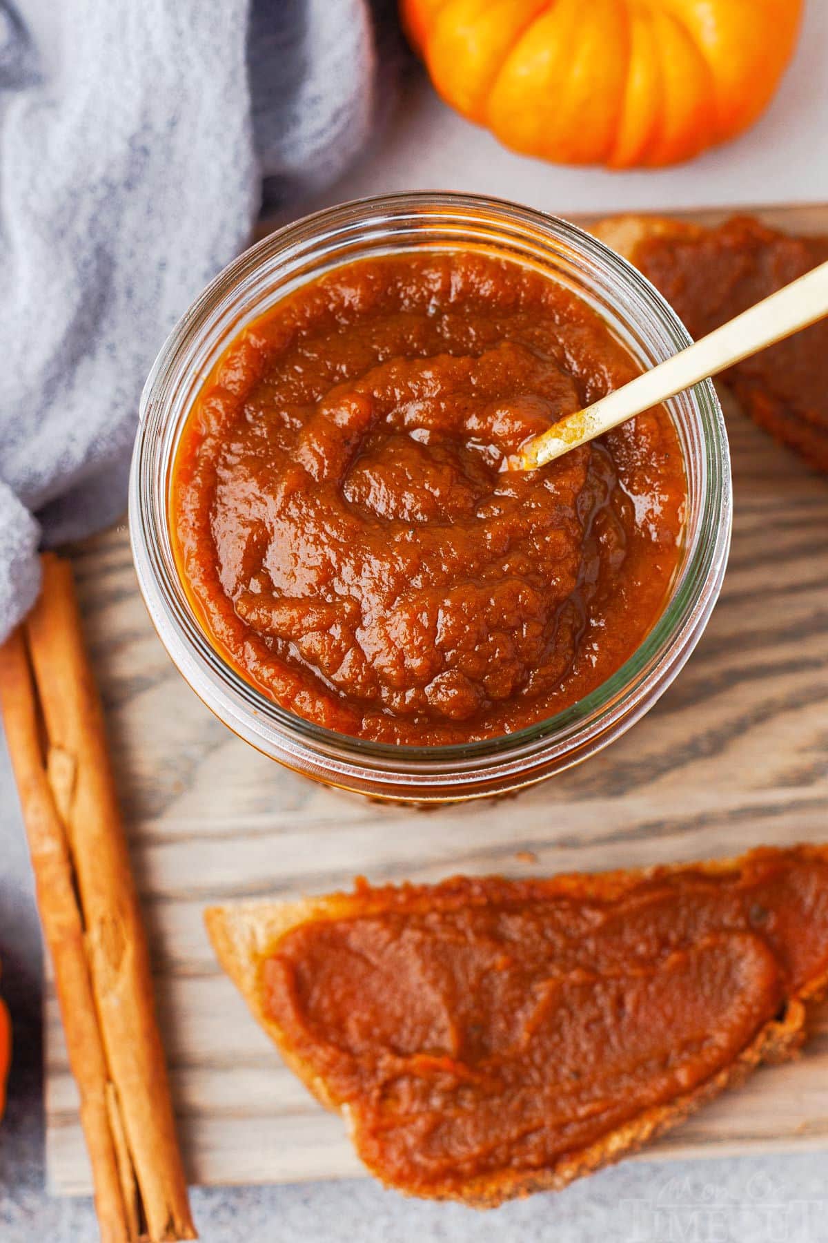 top down view of jar of pumpkin butter with small gold spoon sitting on a gray board with a half piece of toast with pumpkin butter slathered on it.