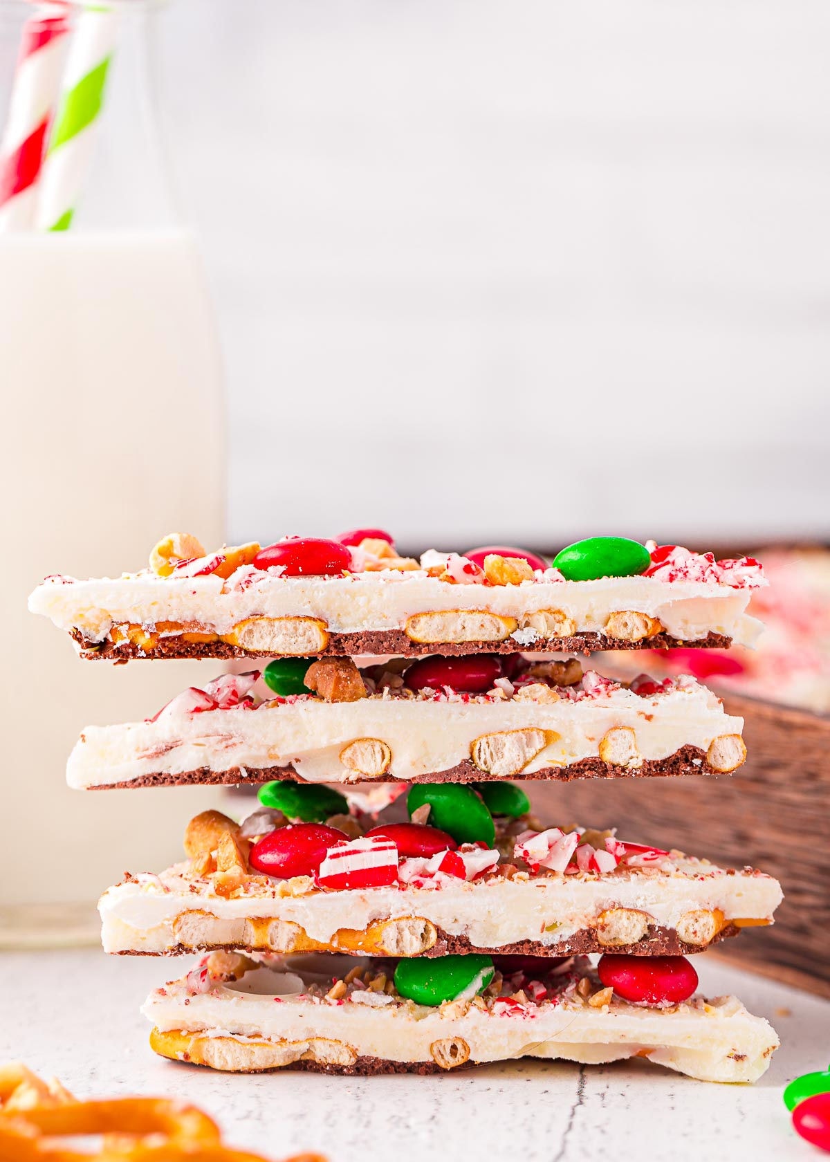 four pieces of christmas bark stacked on top of each other. the bark is made with pretzels, m and ms, and candy canes. glass of milk in background.