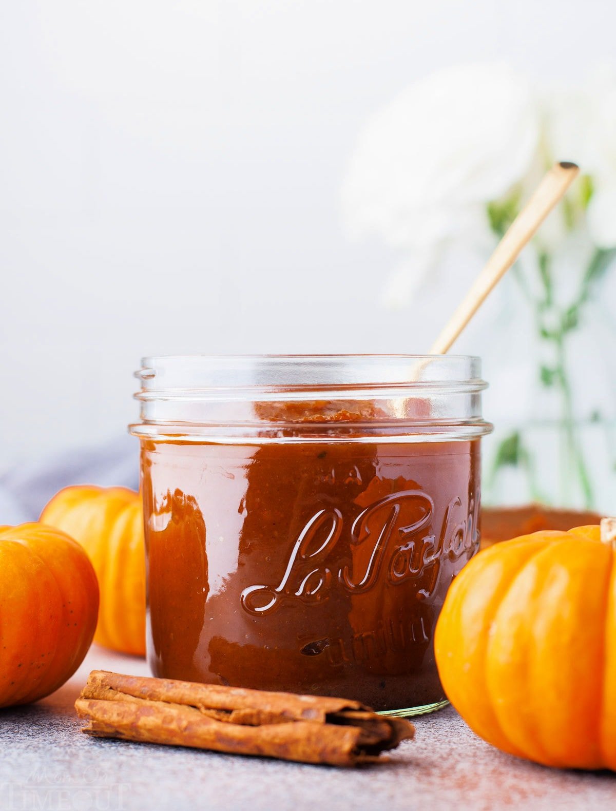 front view of glass jar filled with homemade pumpkin butter. three small decorative pumpkins and a cinnamon stick are sitting around the jar.