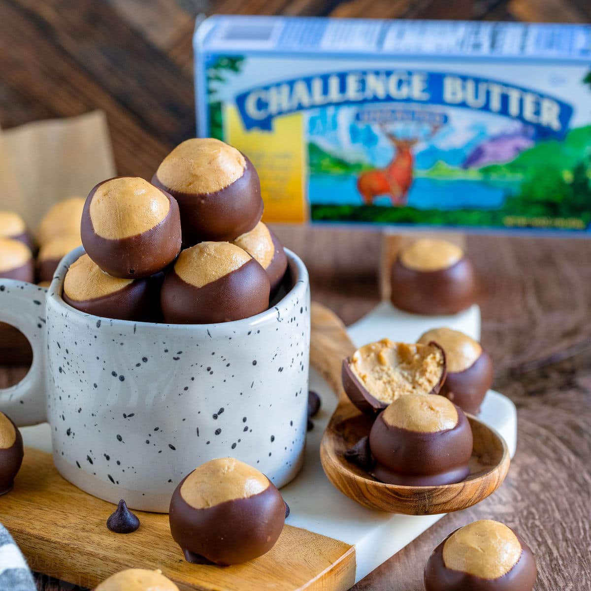 a batch of buckeyes ready to be enjoyed sitting in a white clay mug and next to the mug on a white marble cutting board. a box of challenge butter can be seen in the background.