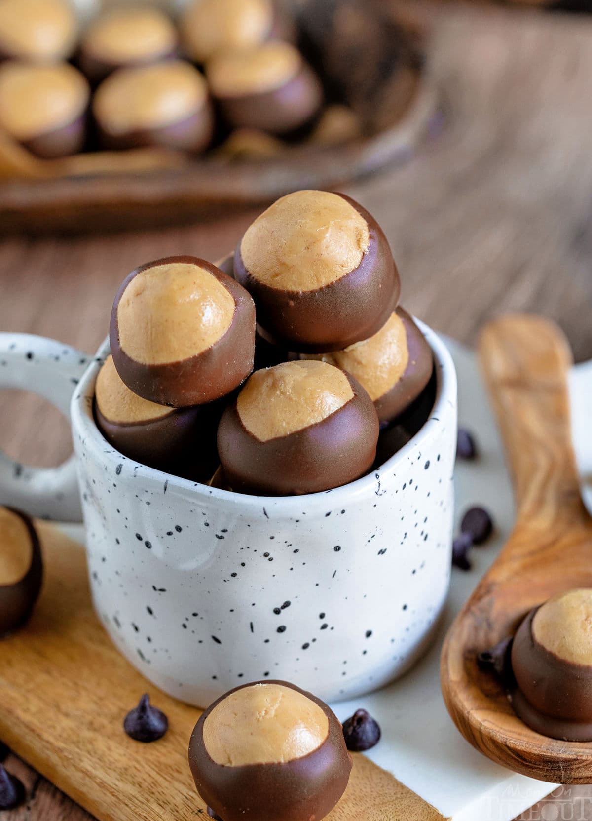 a bunch of buckeye candy filled up a white mug sitting on a white marble surface. more buckeyes can be seen in the background.