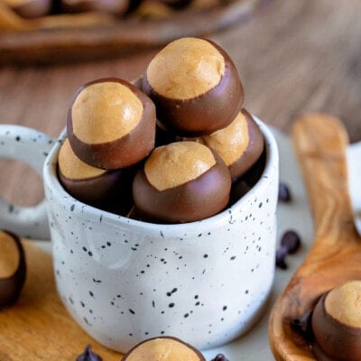 a bunch of buckeye candy filled up a white mug sitting on a white marble surface. more buckeyes can be seen in the background.