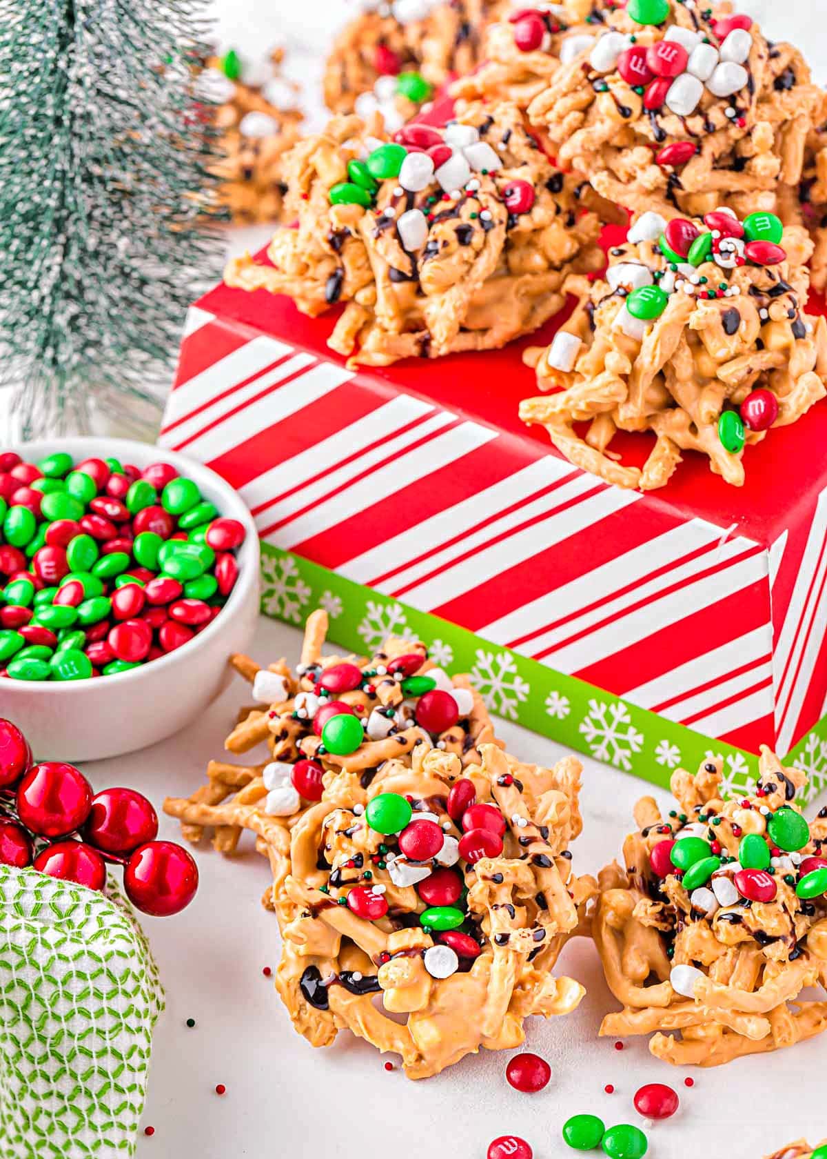 butterscotch haystacks all decorated for christmas sitting on and in front of a red and white striped christmas box. small bowl of red and green m and m candy siting next to the haystacks.