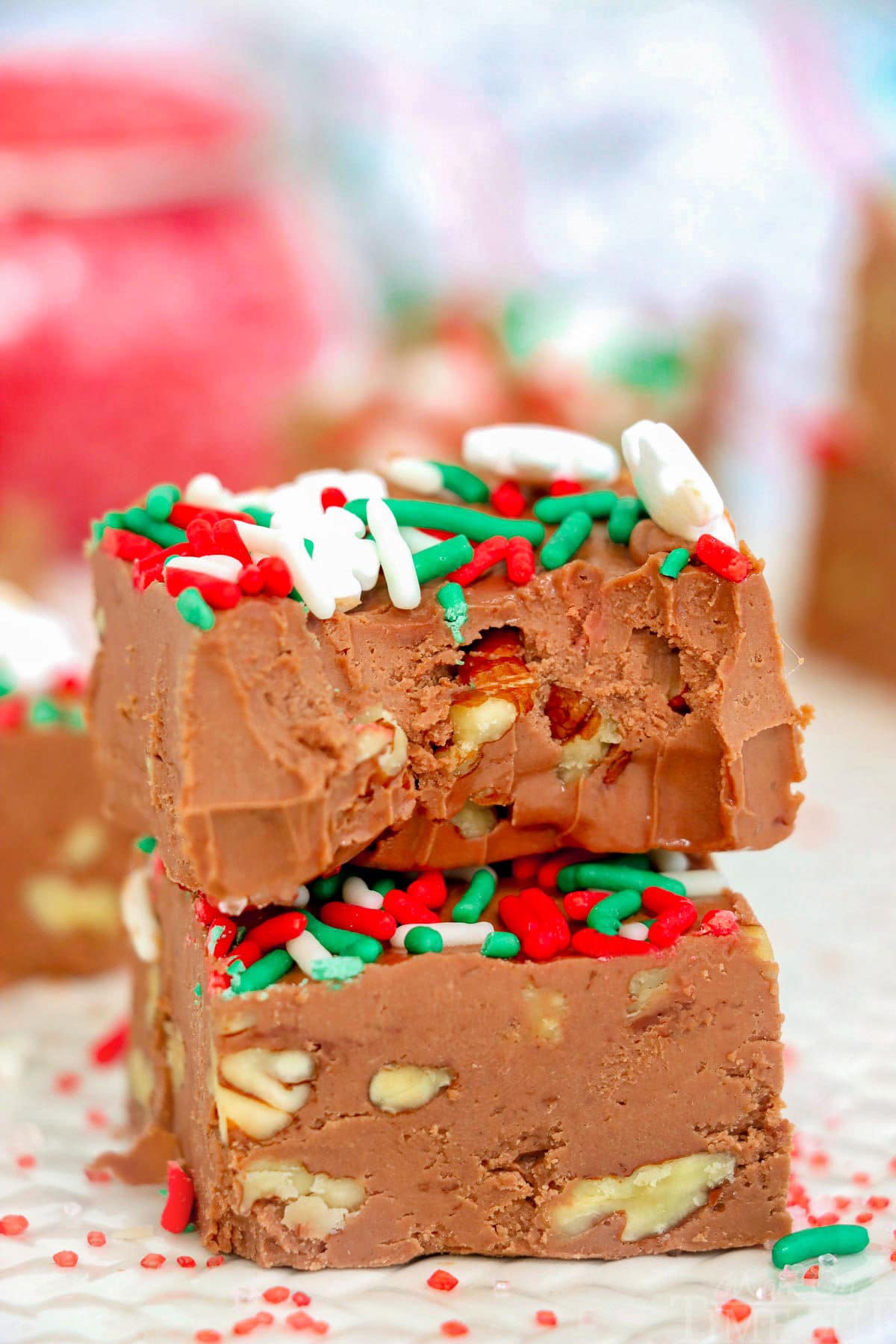 two pieces of chocolate fudge stacked with the top piece having a bite removed. the fudge is topped with christmas sprinkles.