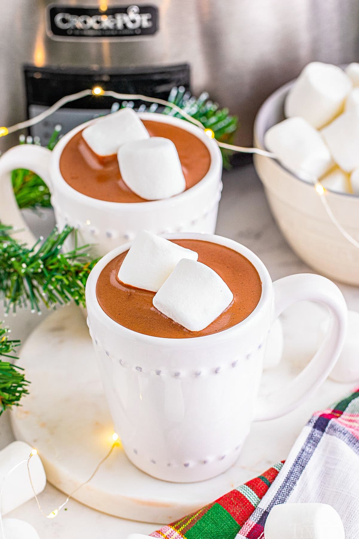 two cups of hot chocolate sitting in front of a silver crockpot and topped with two marshmallows each.