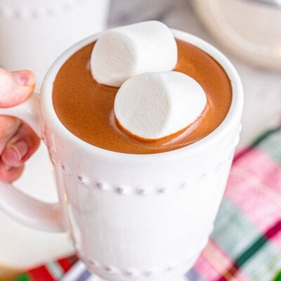 white mug filled with decadent crockpot hot chocolate and topped with two jumbo marshmallows. hand can be seeing holding the mug up and over a christmas linen.