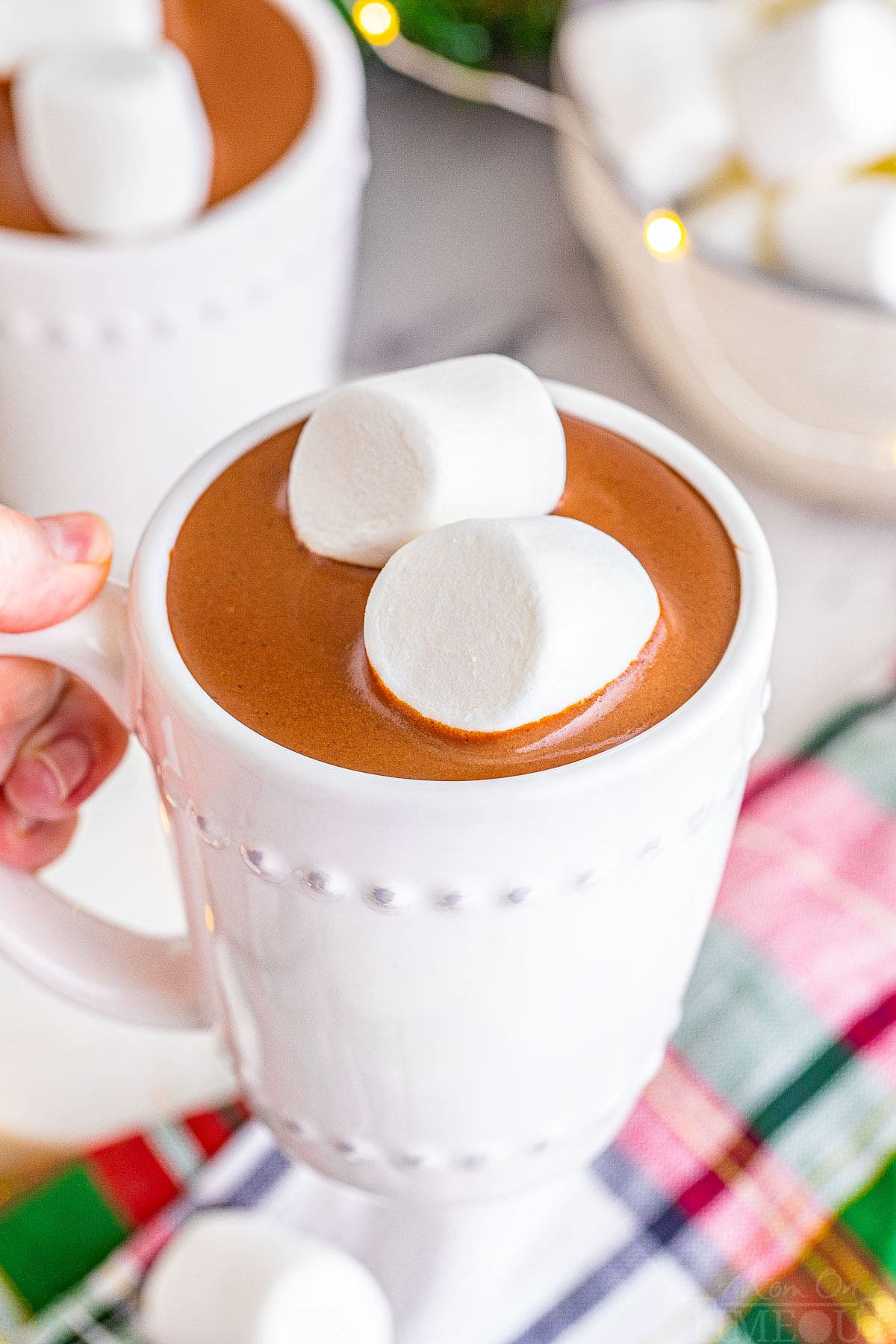 white mug filled with decadent crockpot hot chocolate and topped with two jumbo marshmallows. hand can be seeing holding the mug up and over a christmas linen.