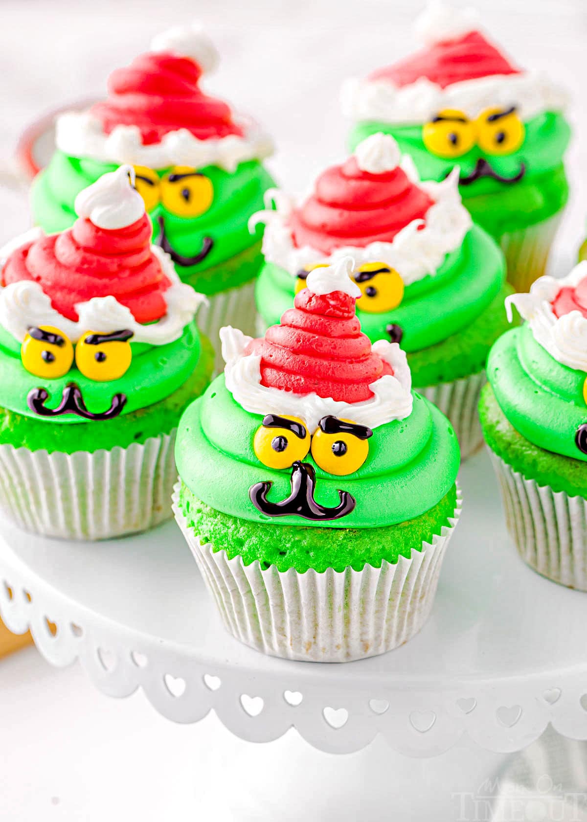 six cupcakes decorated like the grinch with red santa hats sitting on a white cake stand.