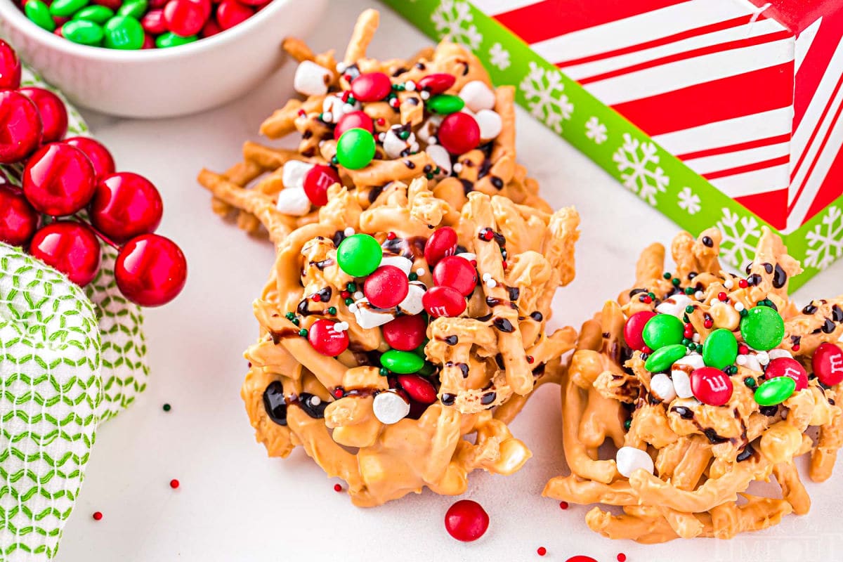 three haystacks candy sitting on white surface in front of a red and white striped christmas box. haystacks are covered with marshmallows and chocolate syrup.