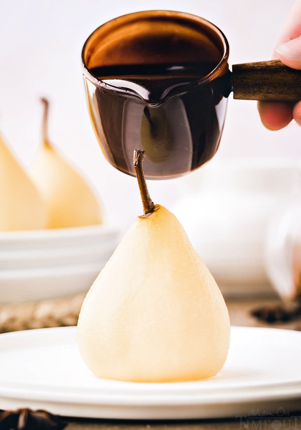 poached pear sitting on white plate with chocolate sauce about to being drizzled onto the pear.