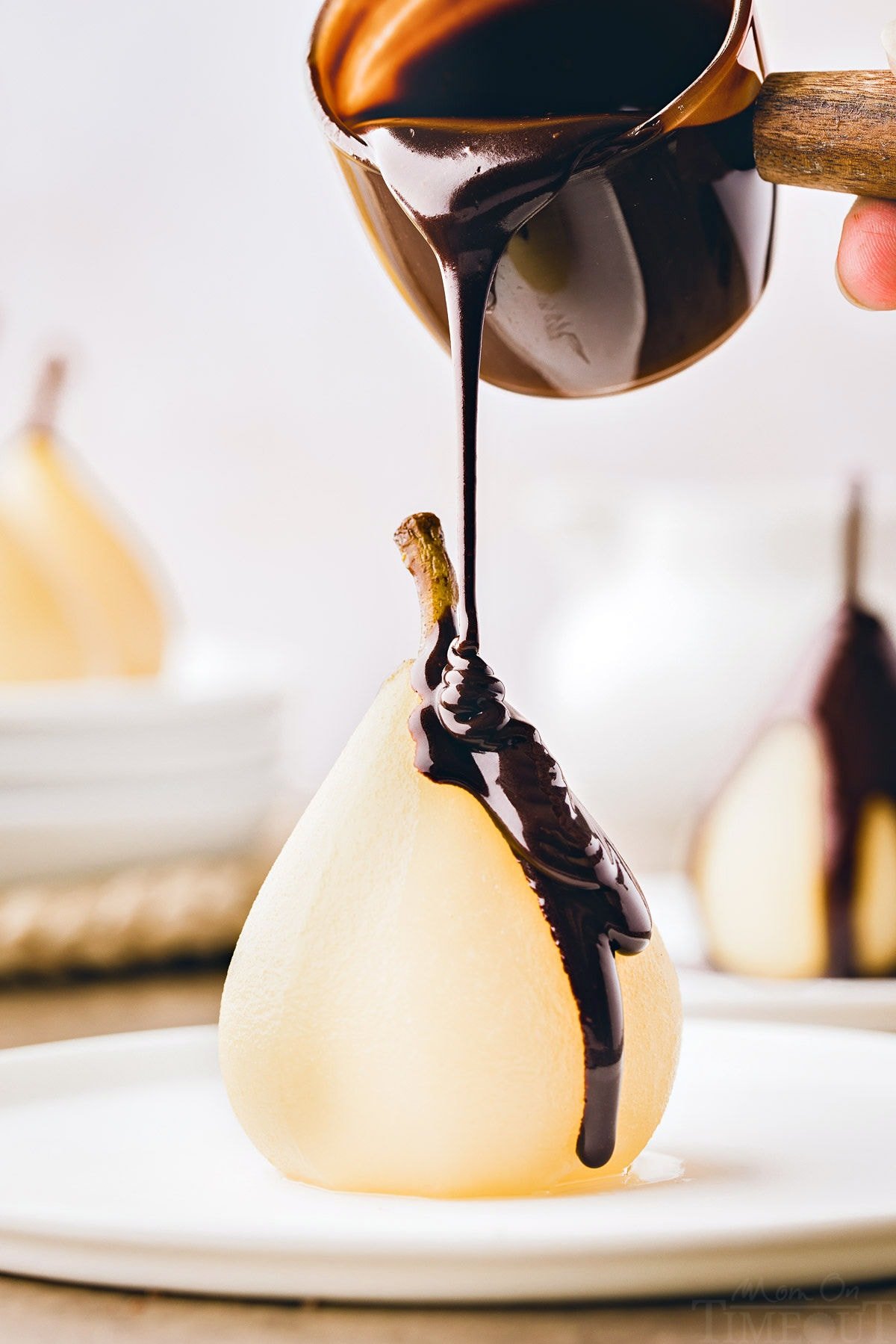 poached pear sitting on white plate with chocolate sauce about to being drizzled onto the pear.