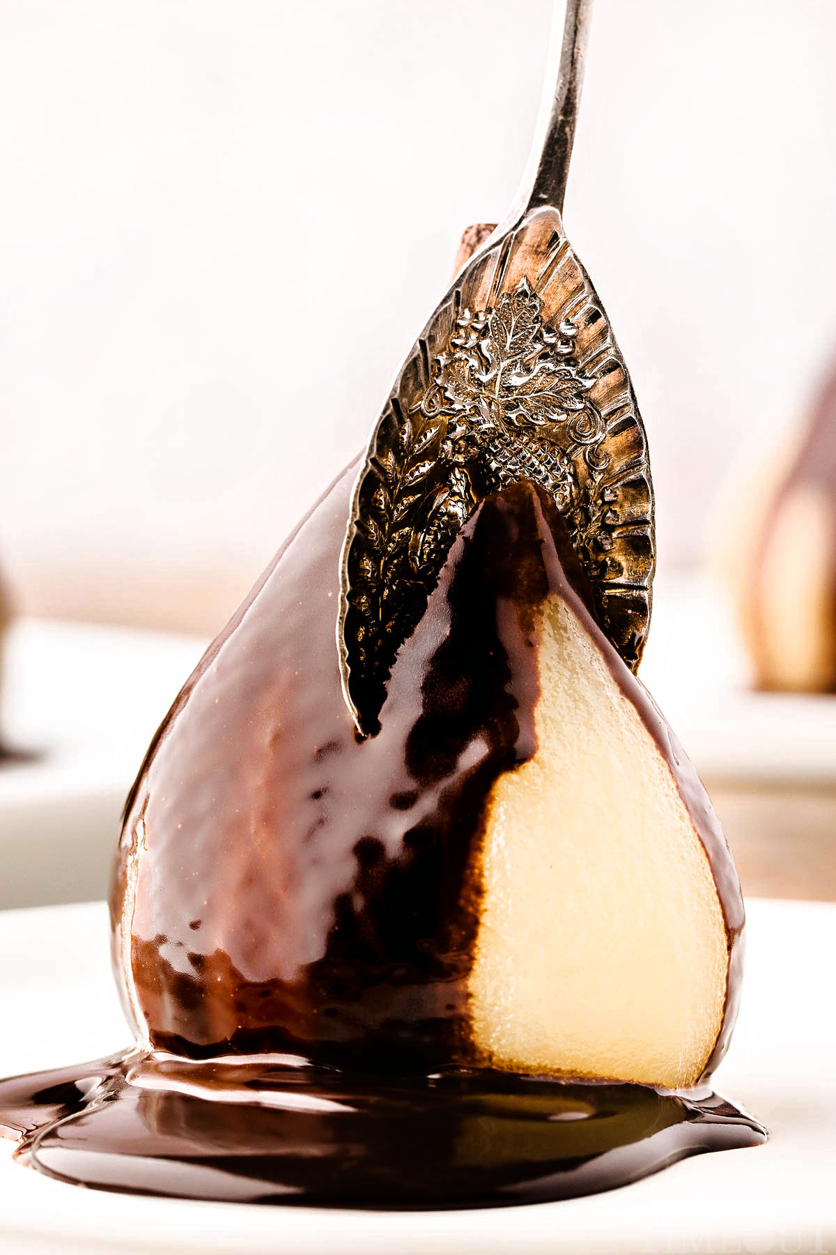 poached pear on white plate topped with chocolate sauce and a vintage spoon is cutting into the pear from the top.