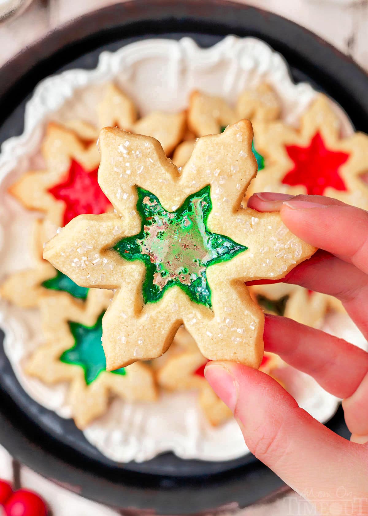 hand holding stained glass cookie over a plate of more cookies. centers are green and red.