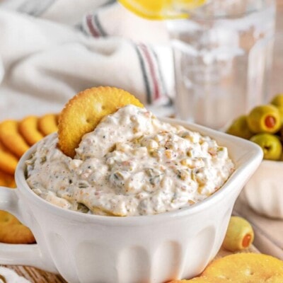 olive dip in a small white bowl sitting on a wood board surrounded by crackers and a small bowl of green olives sitting off to the side. glass of water in the background.