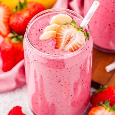 top down angled view of a strawberry banana smoothie in tall glass topped with half a strawberry and banana slices. a pink striped straw is in the smoothie and fresh bananas and strawberries can be seen in the background.