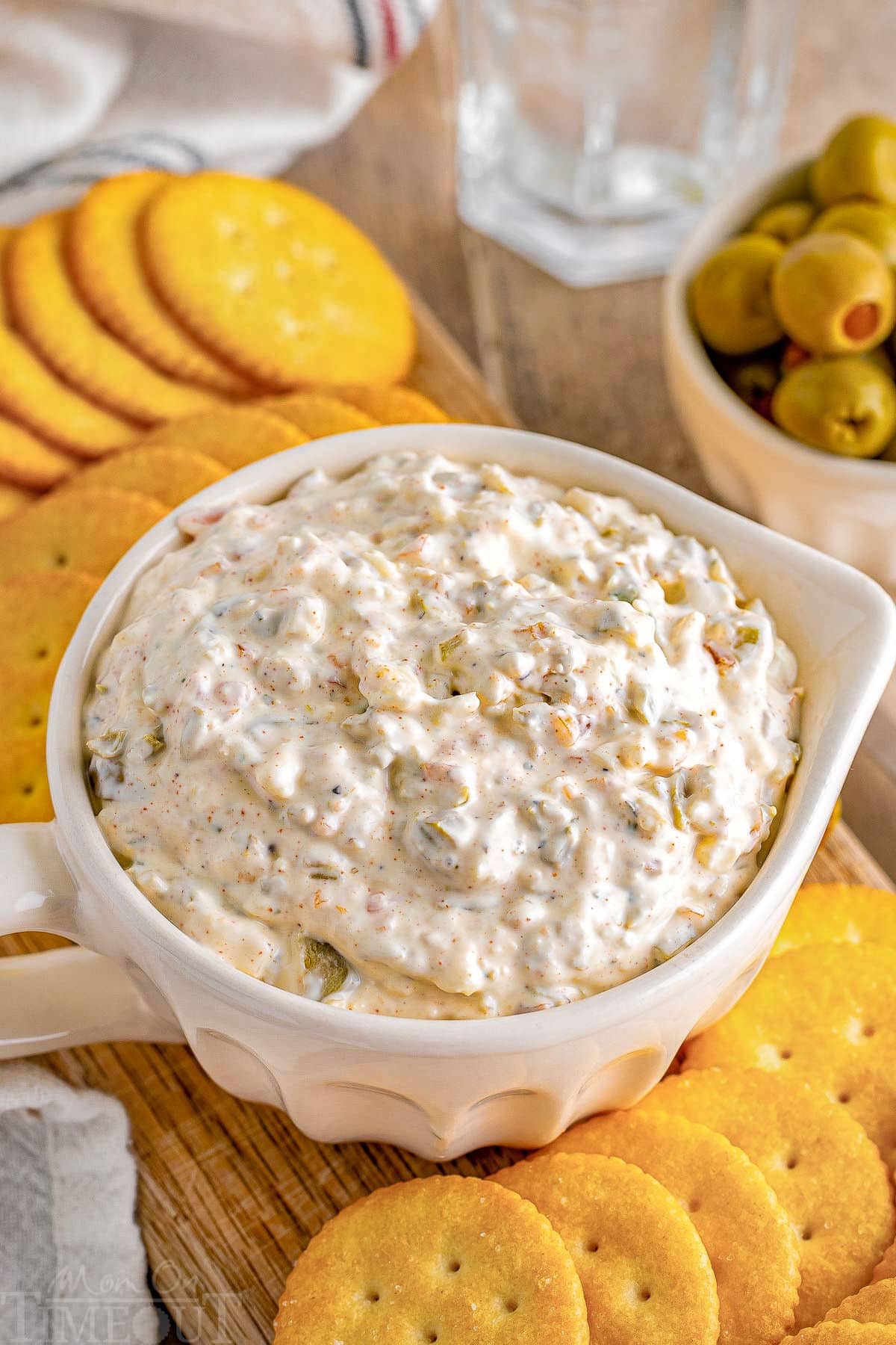top down angled view of green olive dip in a white bowl with a handle. Ritz crackers are on a board next to the dip.
