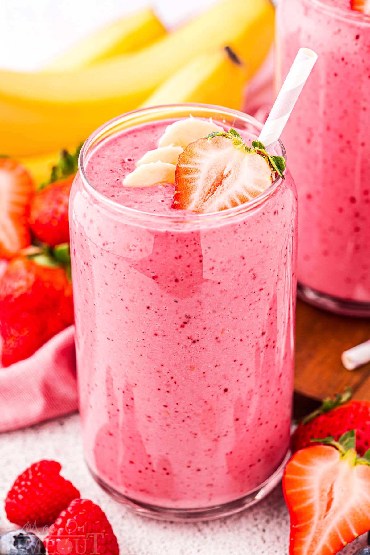 strawberry banana smoothie recipe in a tall glass with a pink and white striped straw. bananas and strawberries can be seen in the background.