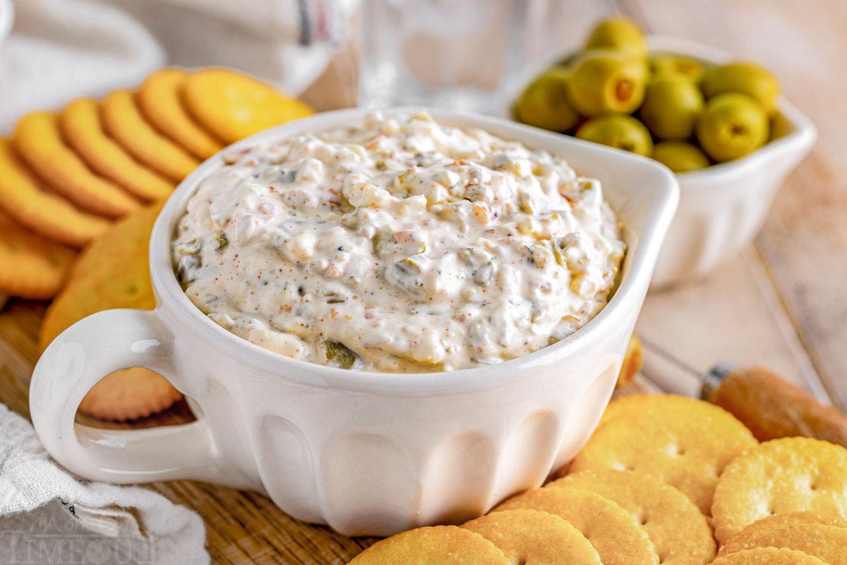 creamy olive dip in a white bowl with crackers surrounding it. a small white bowl with olives can be seen in the background.