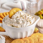 olive dip in a small white bowl sitting on a wood board surrounded by crackers and a small bowl of green olives sitting off to the side. glass of water in the background.