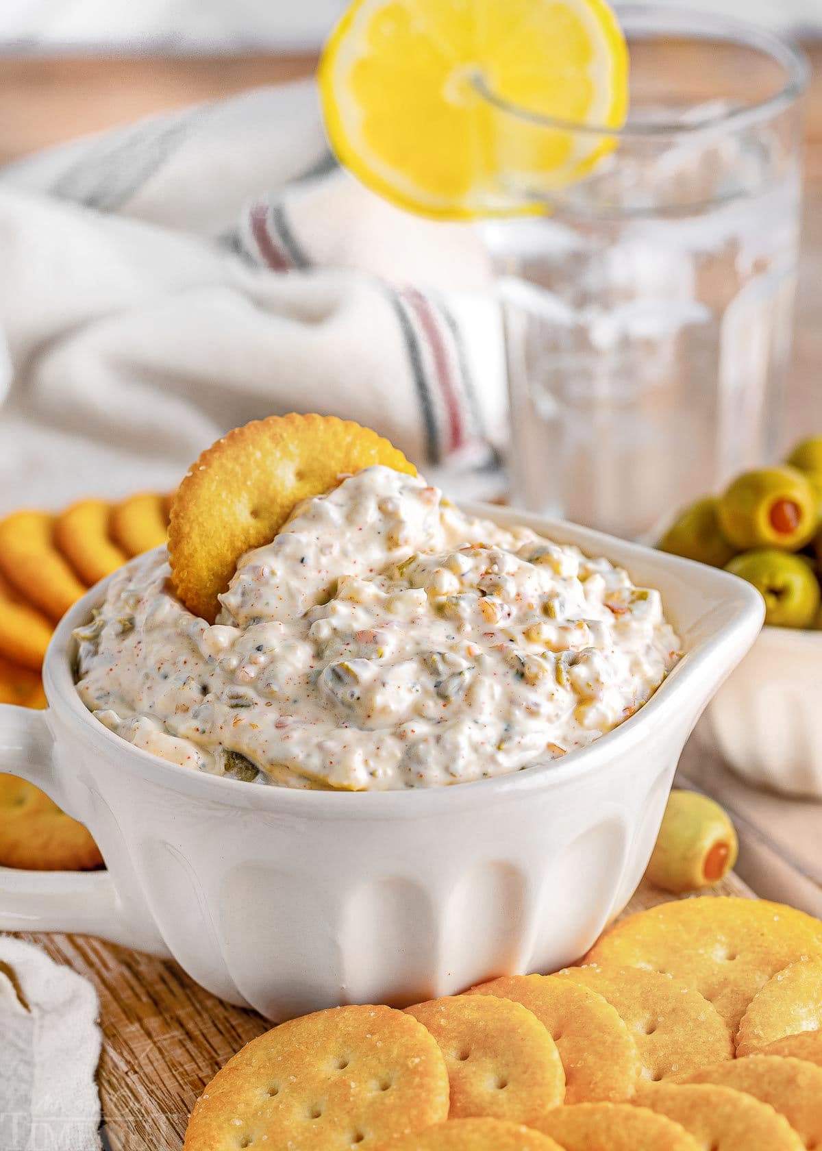 olive dip in a small white bowl sitting on a wood board surrounded by crackers and a small bowl of green olives sitting off to the side. glass of water in the background.