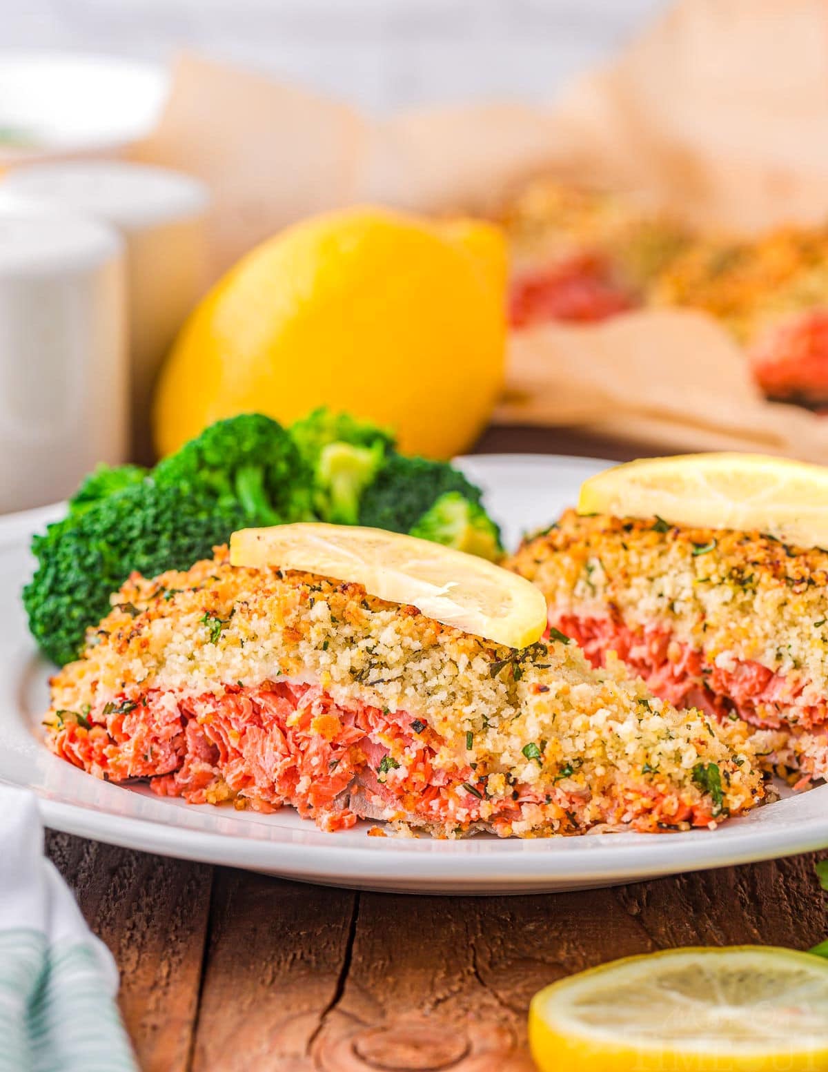 two pieces of salmon on white plate topped with lemon slices and a panko herb crust. lemon and lemon slice can be seen in the background and foreground. 