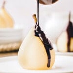 poached pear sitting on white plate with chocolate sauce about to being drizzled onto the pear.