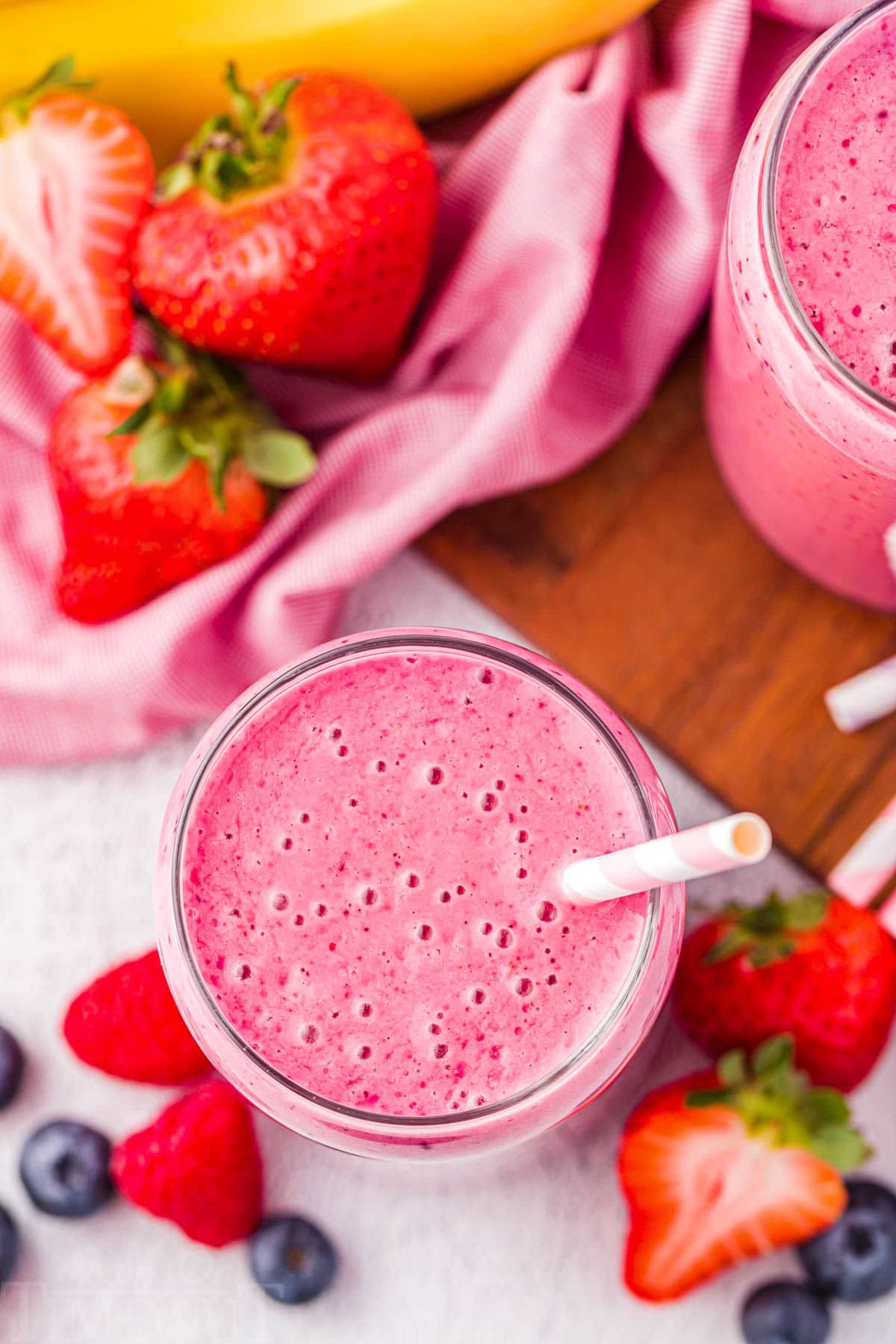 top down vies of the smoothie in a glass cup with a paper straw in it. more strawberries and bananas are scattered by the glass.