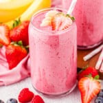 banana strawberry smoothie with a few banana slices and half a strawberry on top. the smoothie is in front of a pink linen and several bananas and strawberries.