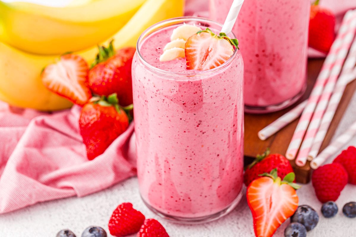 banana strawberry smoothie with a few banana slices and half a strawberry on top. the smoothie is in front of a pink linen and several bananas and strawberries.