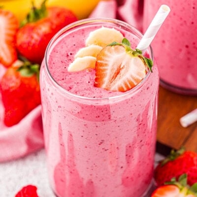 banana strawberry smoothie with a few banana slices and half a strawberry on top. the smoothie is in front of a pink linen and several bananas and strawberries.