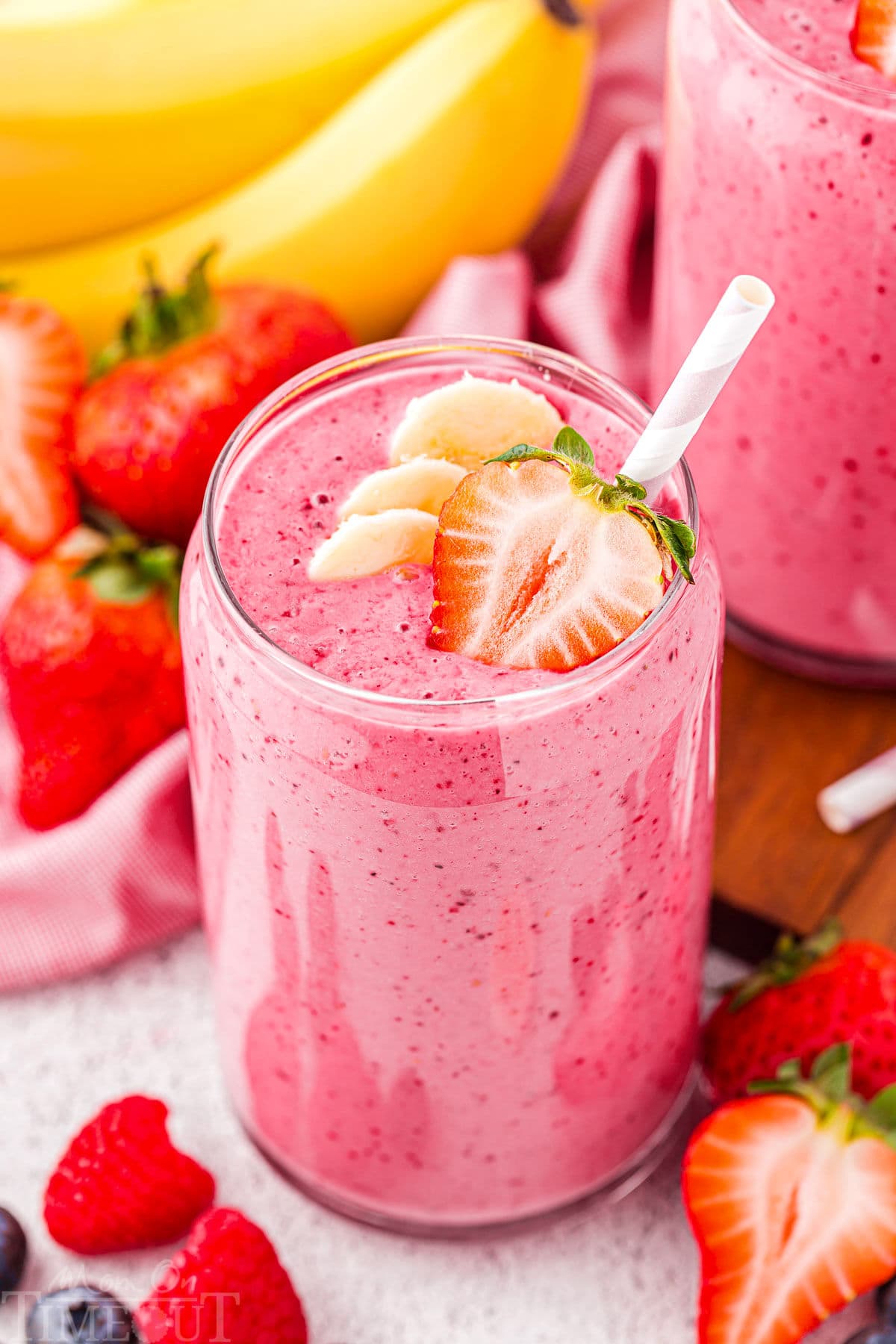 top down angled view of a strawberry banana smoothie in tall glass topped with half a strawberry and banana slices. a pink striped straw is in the smoothie and fresh bananas and strawberries can be seen in the background.