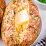 air fryer baked potato on a white plate topped with butter, salt and pepper. Freshly chopped green chives are in a small white bowl in the background.