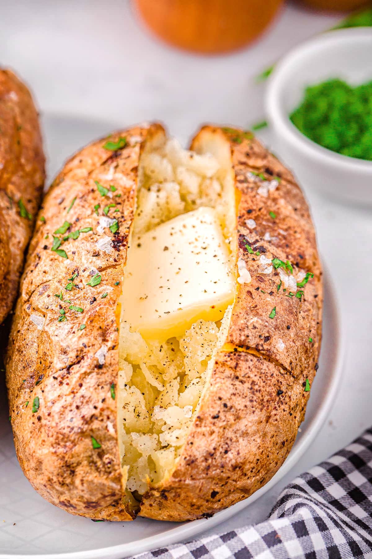 air fryer baked potato on a white plate topped with butter, salt and pepper. Freshly chopped green chives are in a small white bowl in the background.