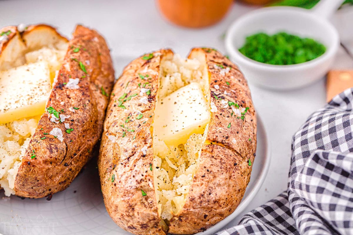 two air fryer baked potatoes on a white plate topped with butter, salt and pepper. Freshly chopped green chives are in a small white bowl in the background.