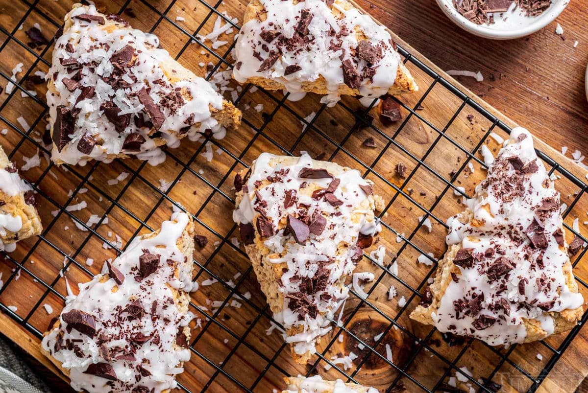 top down look at five scones topped with icing and dark chocolate shavings sitting on a cooling rack on top of a wood board.