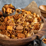 large wooden bowl filled with chex mix with a metal cup in the bowl scooping out the mix.