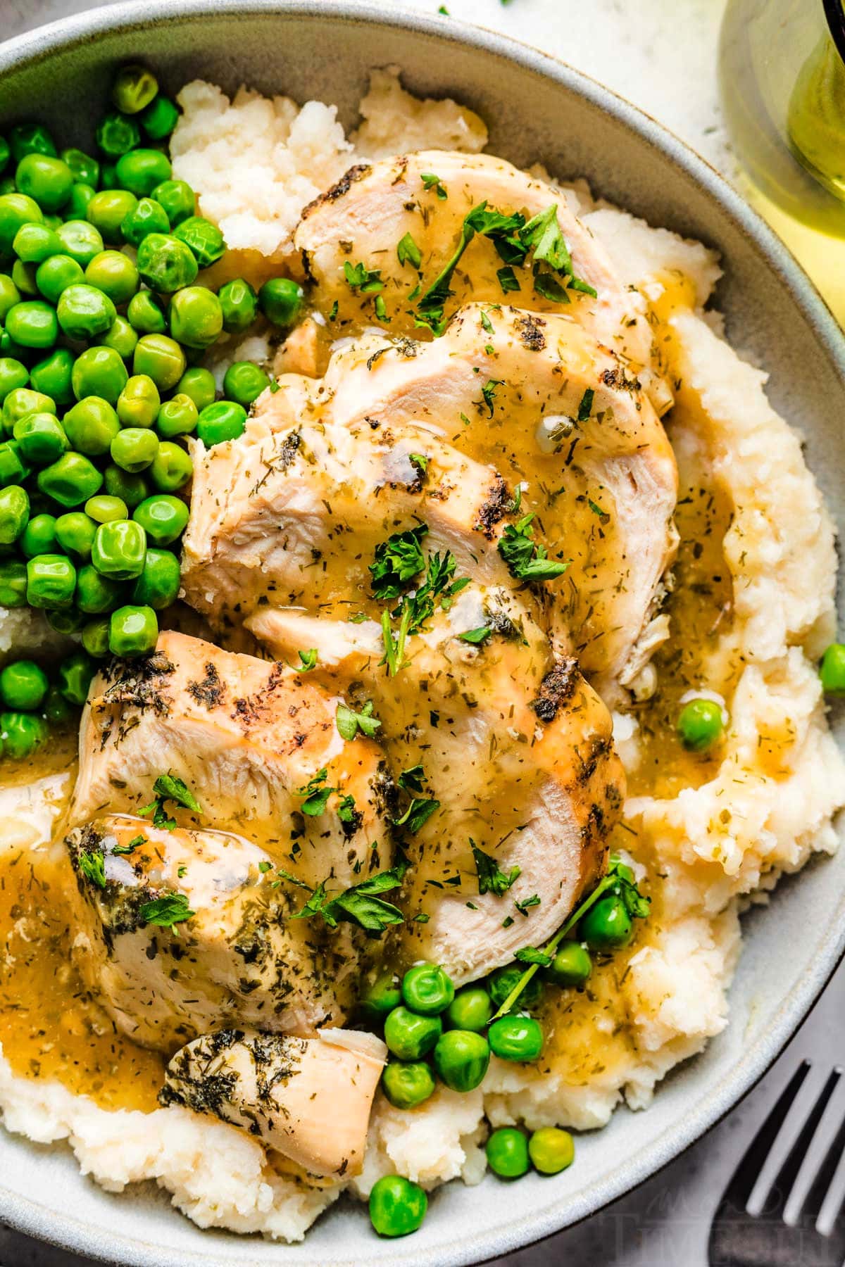 close up of instant pot chicken breast on plate with mashed potatoes and peas and topped with a cream gravy and freshly chopped parsley.